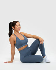 امرأة تجلس على الأرض مرتدية حمالة صدر نسائية من BEST Define Seamless Racer Back باللون الأزرق الدخاني وسروالًا ضيقًا، مع التركيز على أحزمة الظهر المتقاطعة.