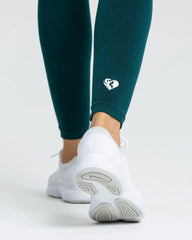 أرجل امرأة ترتدي طماق Move Seamless | Smaragd Green Solid من WOMEN'S BEST وحذاء رياضي أبيض.