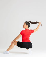 امرأة ترتدي تي شيرت BEST Power Seamless للنساء باللون الأحمر وتؤدي تمرين القرفصاء على خلفية بيضاء، وتظهر قوتها والنسيج عالي الجودة للملابس.