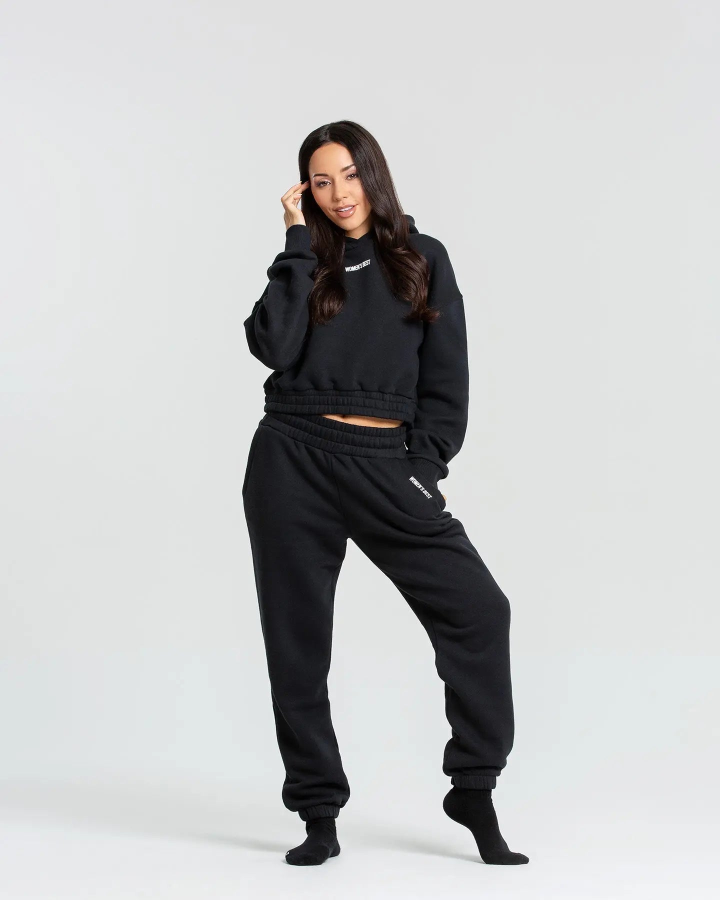 وصف: امرأة ترتدي كنزة قلنسوة Comfort Cropped Hoodie أسود وسروال رياضي أسود من WOMEN'S BEST.