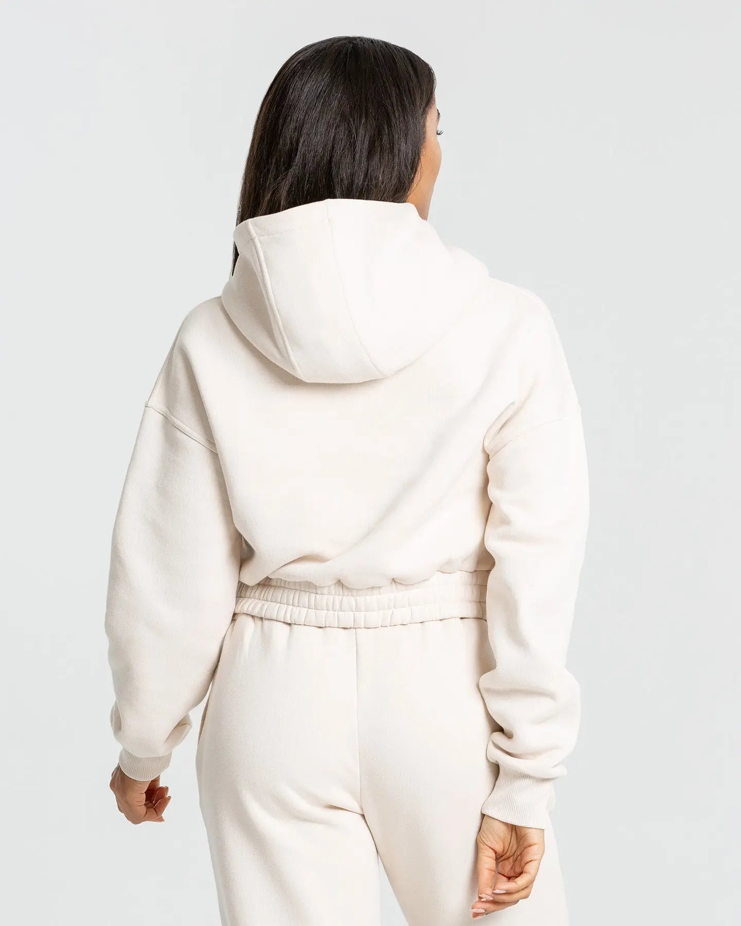 ترتدي الصورة الخلفية لامرأة بغطاء للرأس Comfort Cropped Hoodie بلون كريم وسروال ريا. العلامة التجارية أفضل النساء