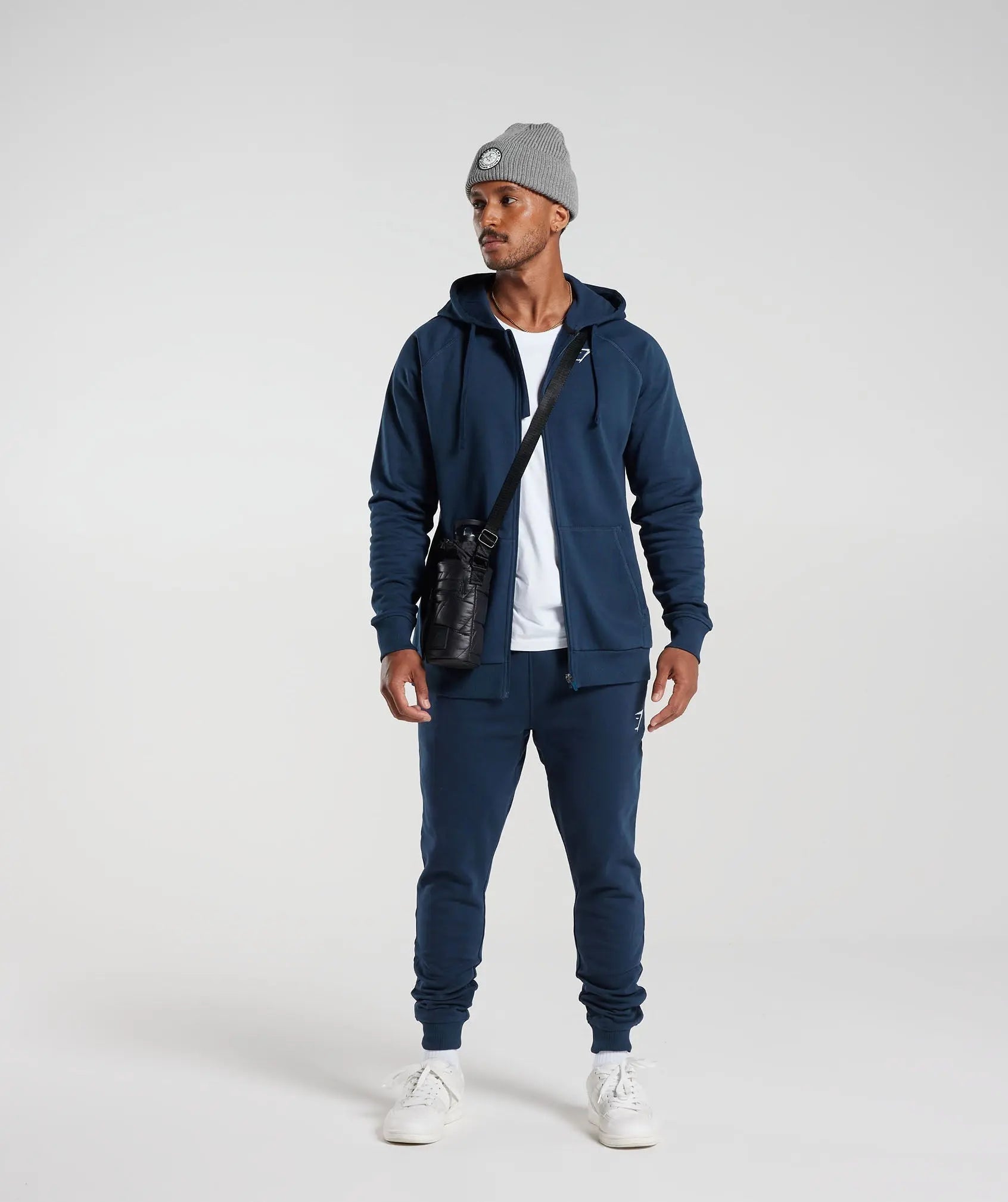 يرتدي الرجل هودي Crest Zip Up باللون الأزرق الداكن من Gymshark | United We Sweat وقبعة رمادية.