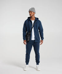 يرتدي الرجل هودي Crest Zip Up باللون الأزرق الداكن من Gymshark | United We Sweat وقبعة رمادية.