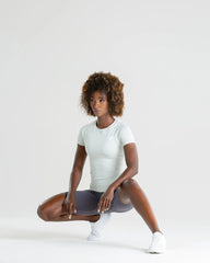 امرأة سوداء تجلس القرفصاء على خلفية بيضاء، وترتدي تي شيرت BEST Power Seamless للسيدات باللون الرمادي الفاتح المغسول.